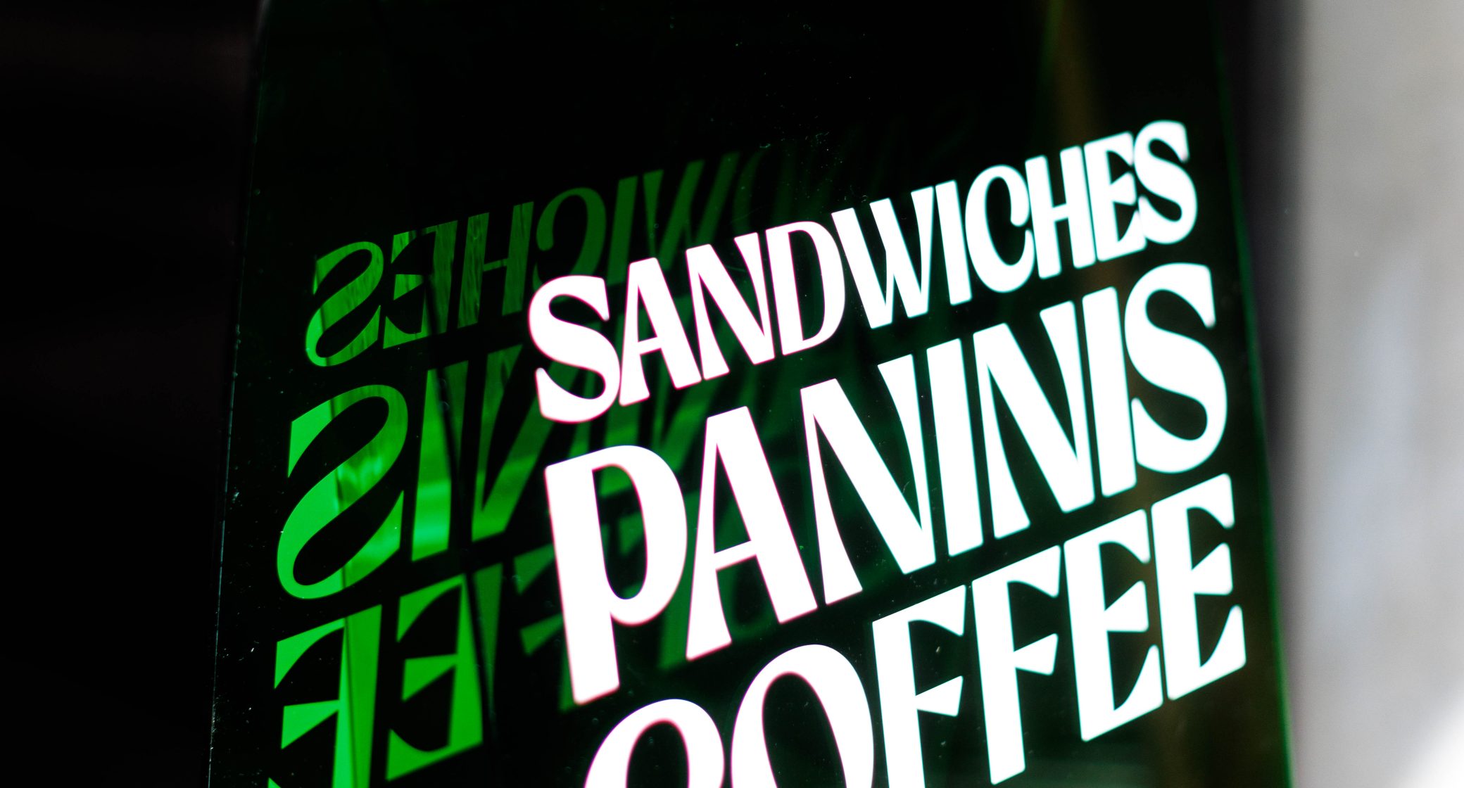 A clear sign displays the words "SANDWICHES," "PANINIS," and "COFFEE," with a right-pointing arrow, against a blurred background.