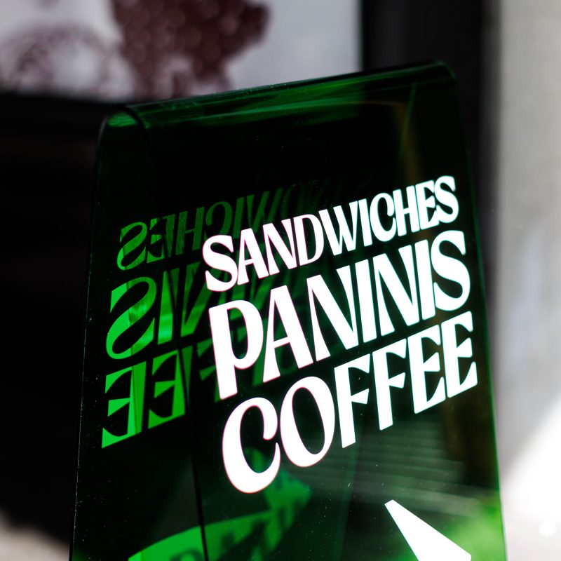 A clear sign displays the words "SANDWICHES," "PANINIS," and "COFFEE," with a right-pointing arrow, against a blurred background.