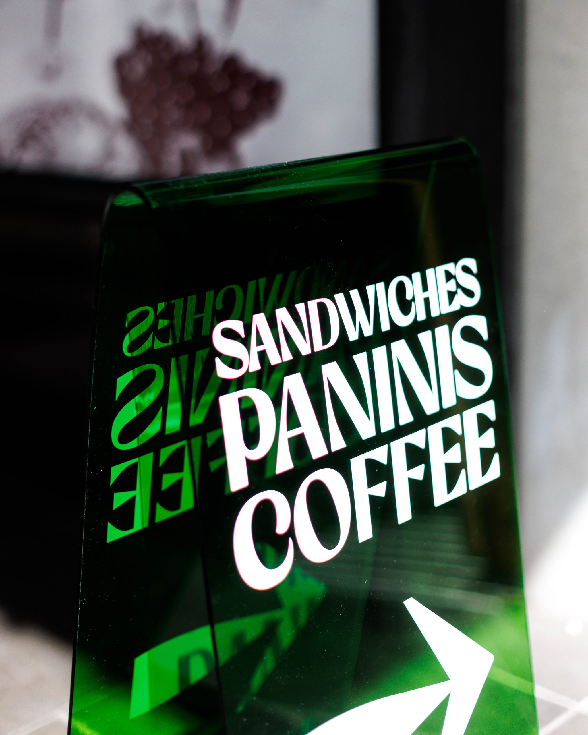 A clear sign displays the words "SANDWICHES," "PANINIS," and "COFFEE," with a right-pointing arrow, against a blurred background.