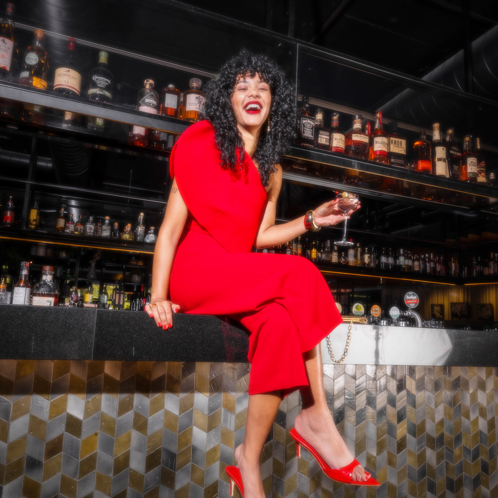 A woman in a bright red dress and heels sits at a bar, laughing and holding a stemmed glass. Liquor bottles line the shelves behind her.