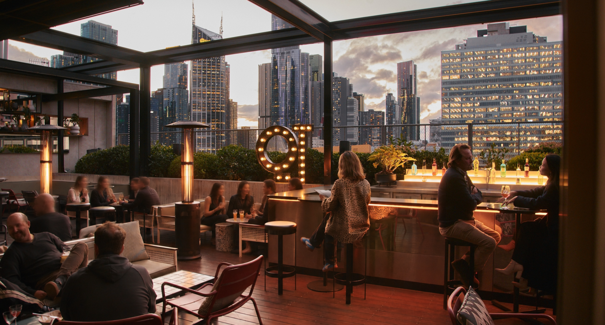 A rooftop bar setting during sunset, featuring patrons seated at tables and the city skyline in the background.