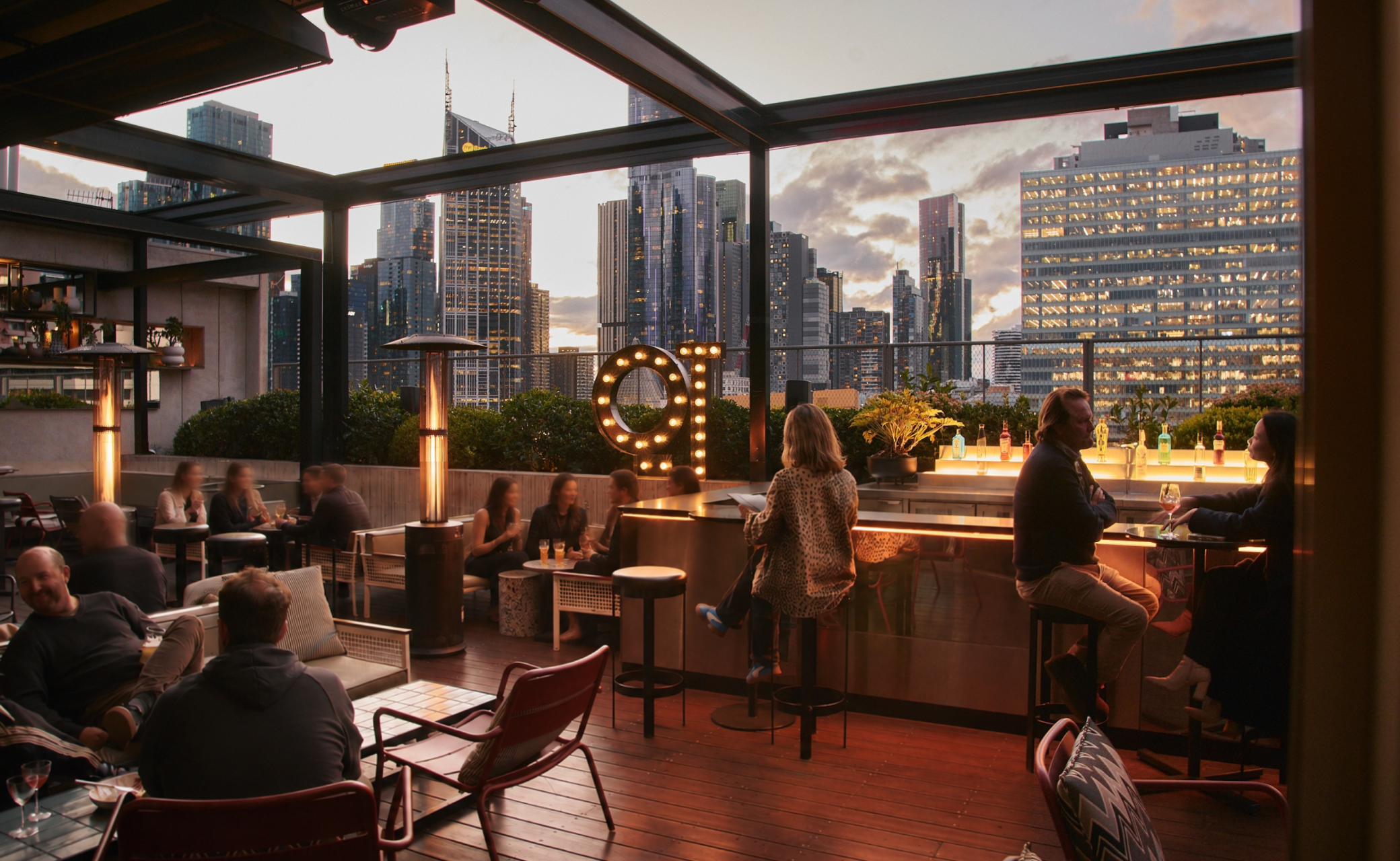 A rooftop bar setting during sunset, featuring patrons seated at tables and the city skyline in the background.