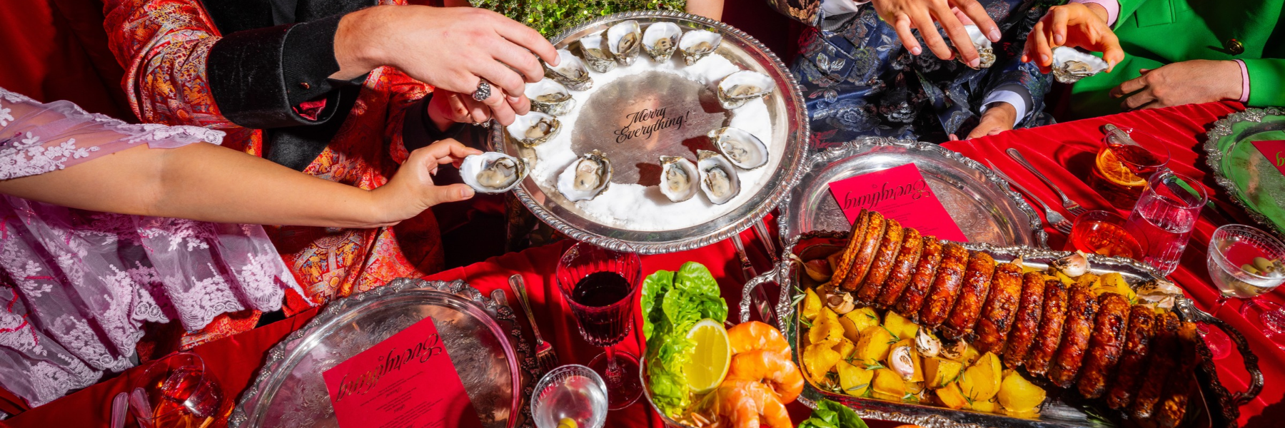 Hands reach for oysters on a silver platter beside roasted meat, seafood, and drinks on a red-covered banquet table.