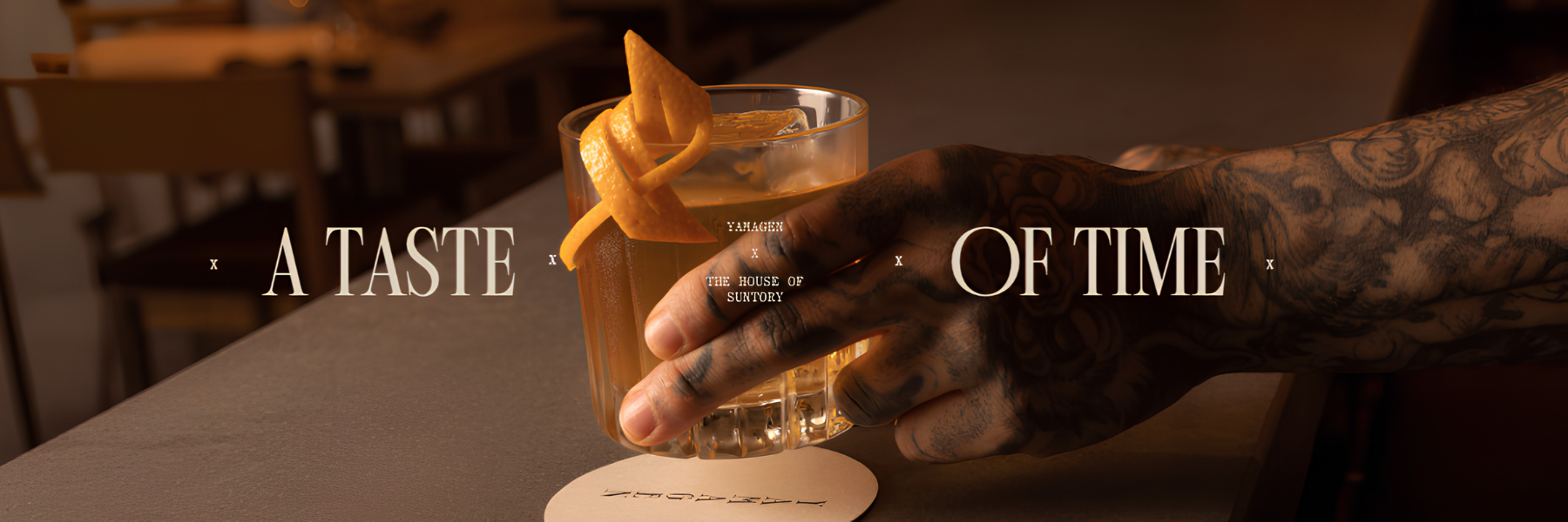 A tattooed hand holds an amber cocktail with an orange twist above a coaster, beneath the words “A Taste of Time.”