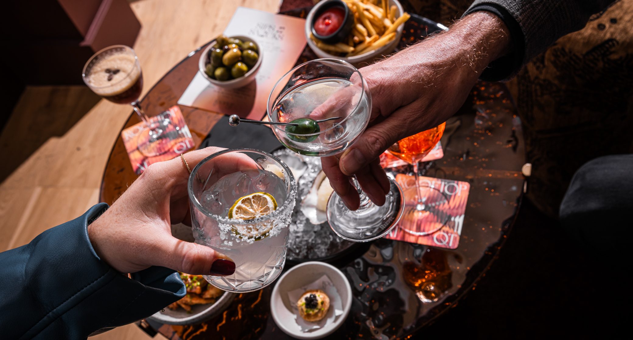 Two people hold cocktails over a table with olives, fries, and small plates of food.