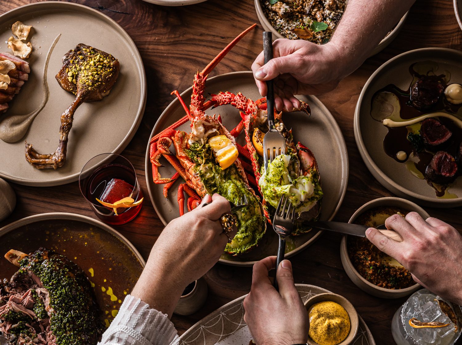 Overhead view of diners sharing a lavish meal, cutting lobster and assorted meats at a wooden table with wine glasses.