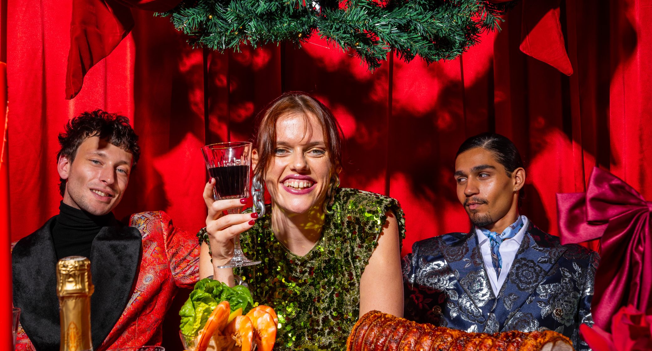 Three elegantly dressed individuals sit at a festive table adorned with seafood, floral arrangements, and champagne, against a vibrant red backdrop.