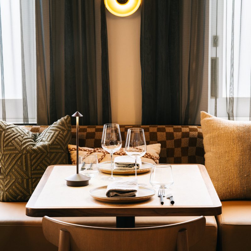 A cozy restaurant setting features a well-set table with glasses and plates, flanked by patterned pillows on a cushioned banquette. Soft lighting and sheer curtains enhance the ambiance.