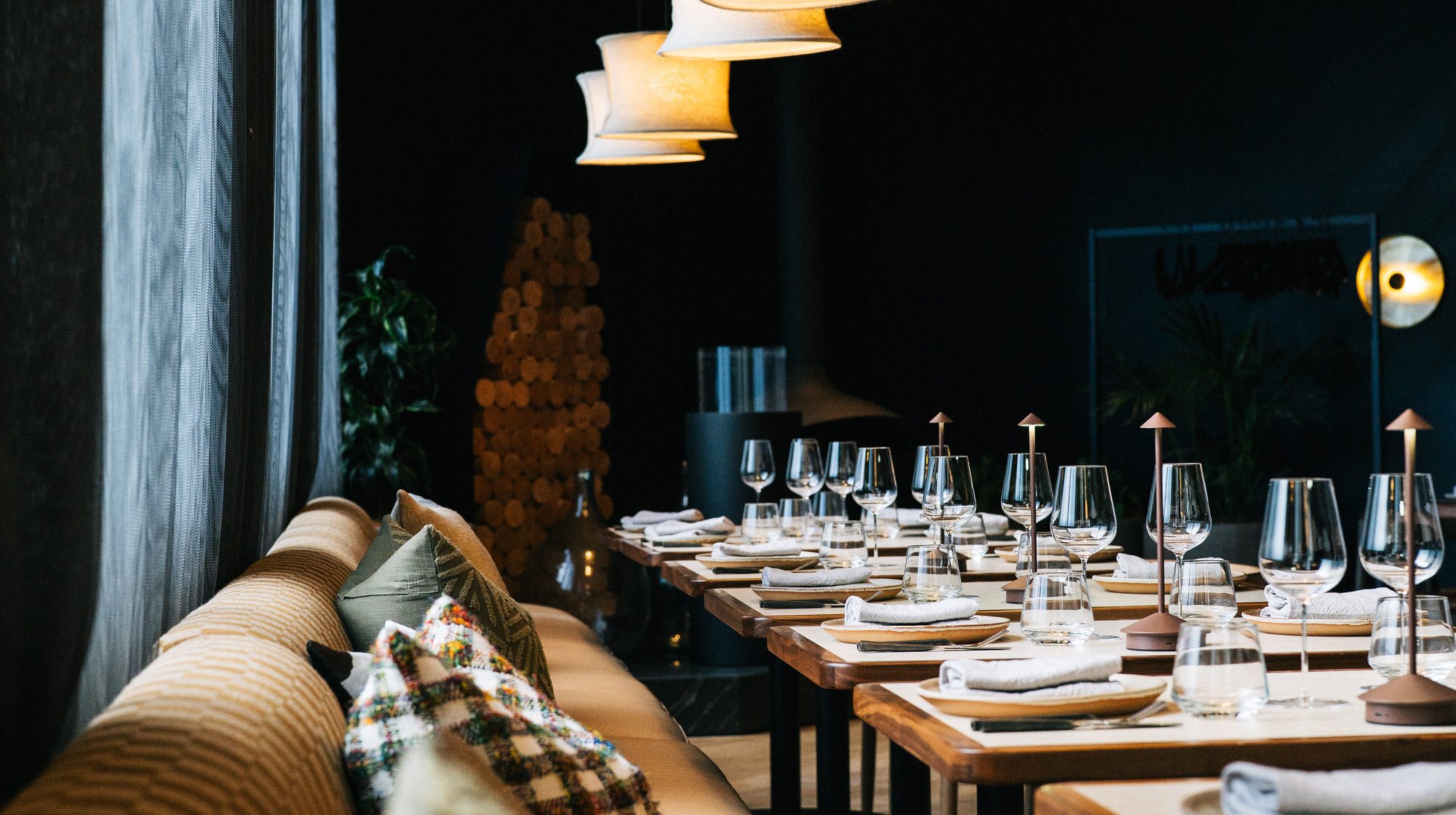 A dim restaurant dining room with set tables, wine glasses, cushioned bench seating, and glowing pendant lamps.