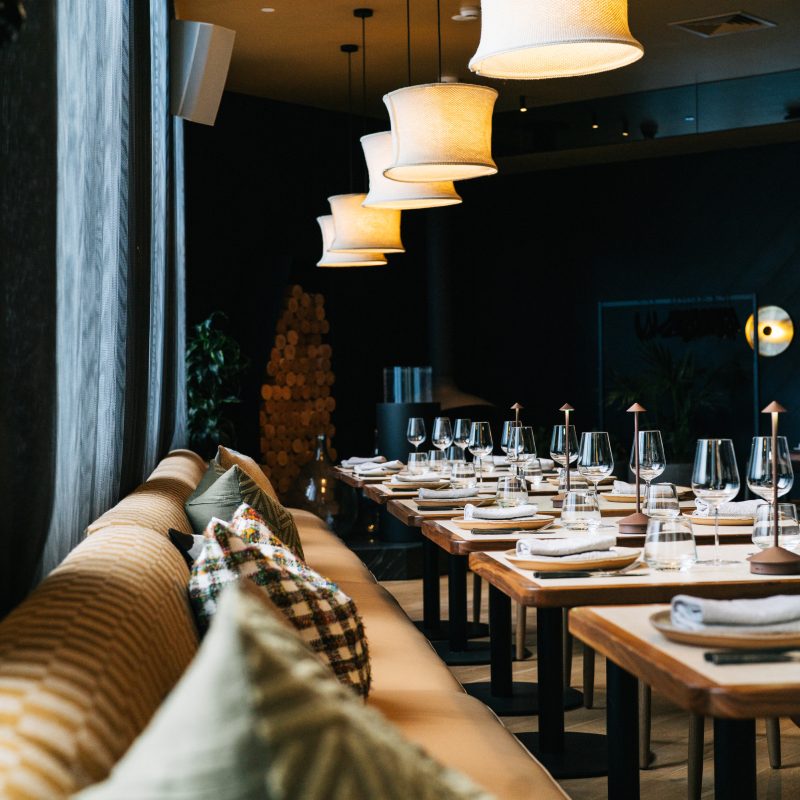 A dim restaurant dining room with set tables, wine glasses, cushioned bench seating, and glowing pendant lamps.