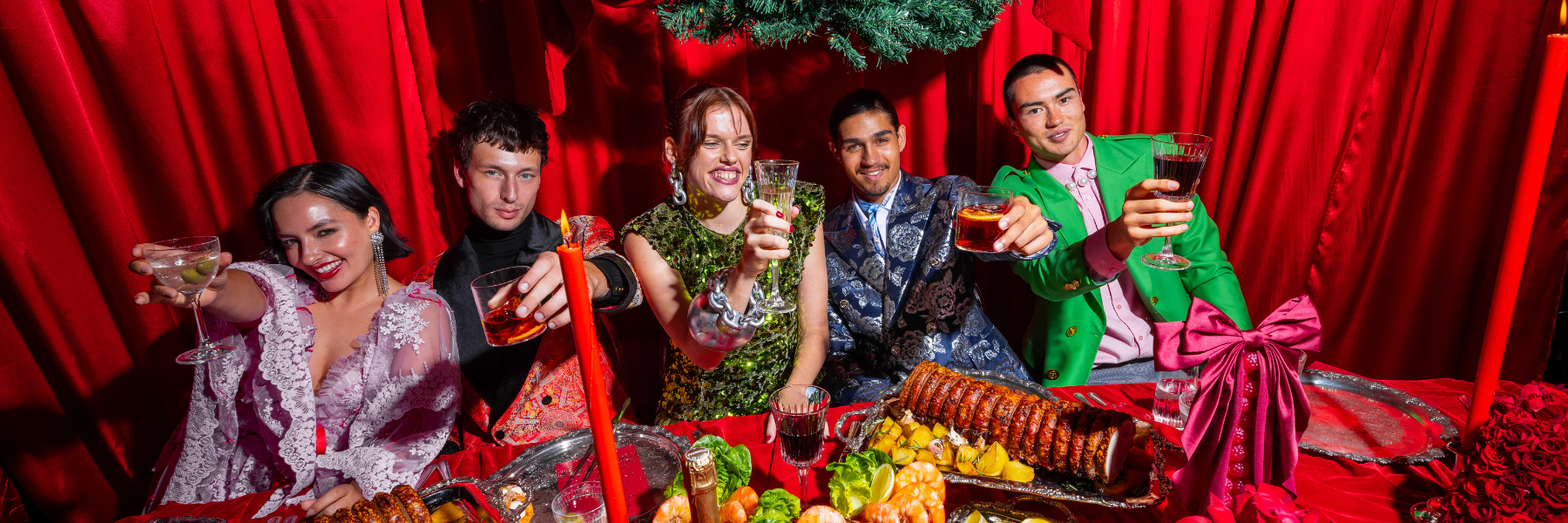 A group of five people toast with drinks at a festive table adorned with food, candles, and a red backdrop.