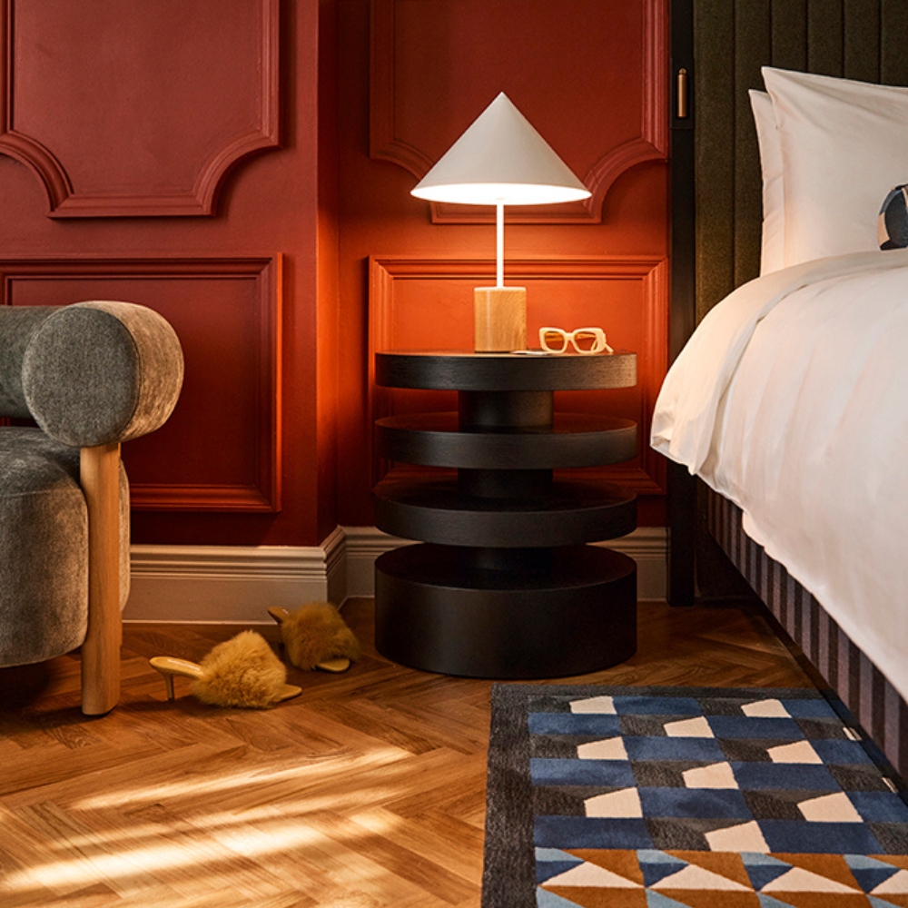 Bedroom corner with a white bed, layered round black bedside table and lamp, gray chair, slippers, and geometric rug against red paneled walls.