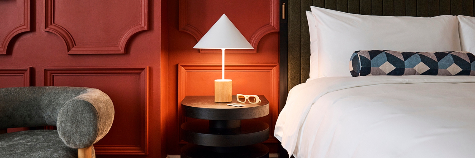 White bed with a geometric bolster beside a round nightstand and lamp, against red paneled walls; gray chair at left.