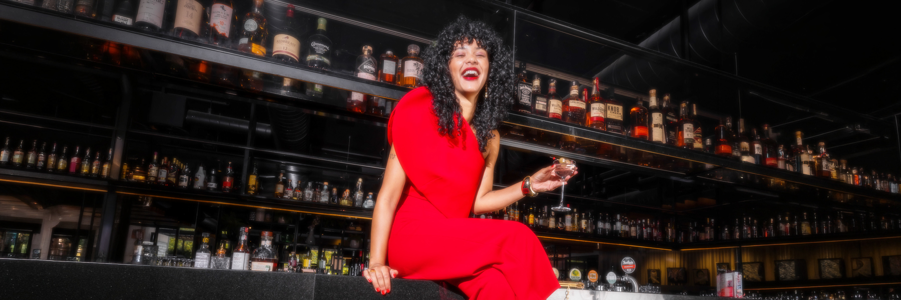 A woman in a bright red dress sits at a bar, holding a stemmed glass in front of shelves of liquor bottles.