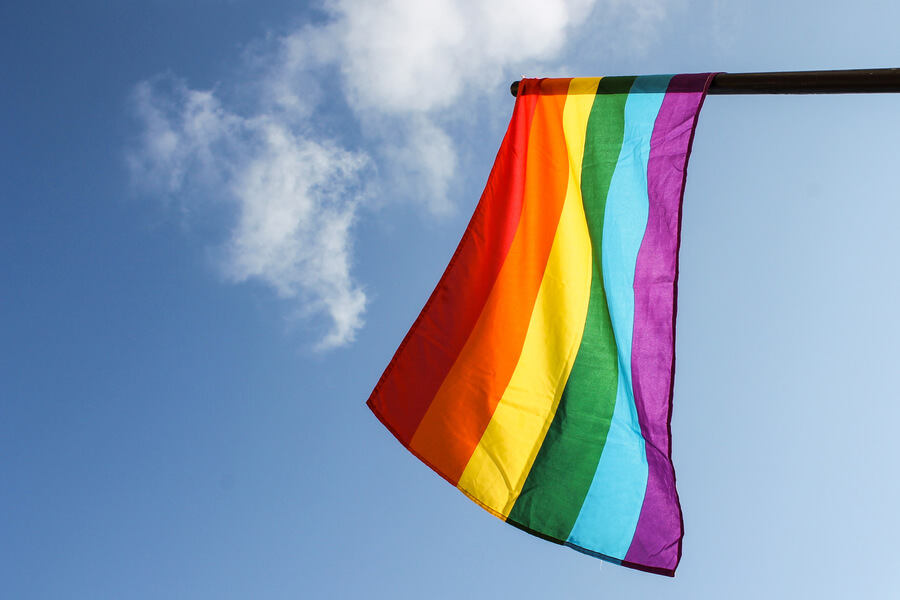 A rainbow flag waves against a clear blue sky with a few clouds. The flag features horizontal stripes in red, orange, yellow, green, blue, and purple.