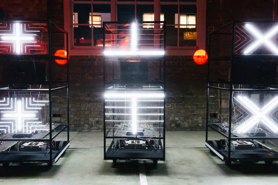 Three illuminated structures with geometric patterns, including crosses and X shapes, are displayed in a dimly lit space.