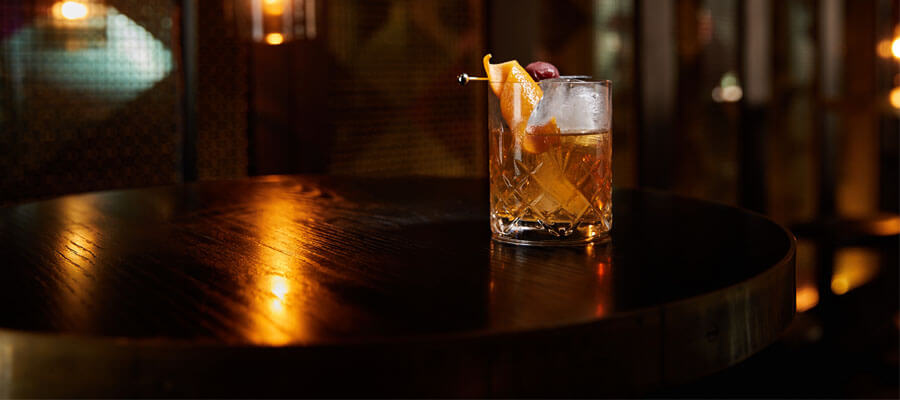 A whiskey cocktail with an orange slice and cherry sits on a dark wooden table, illuminated by warm ambient lights.