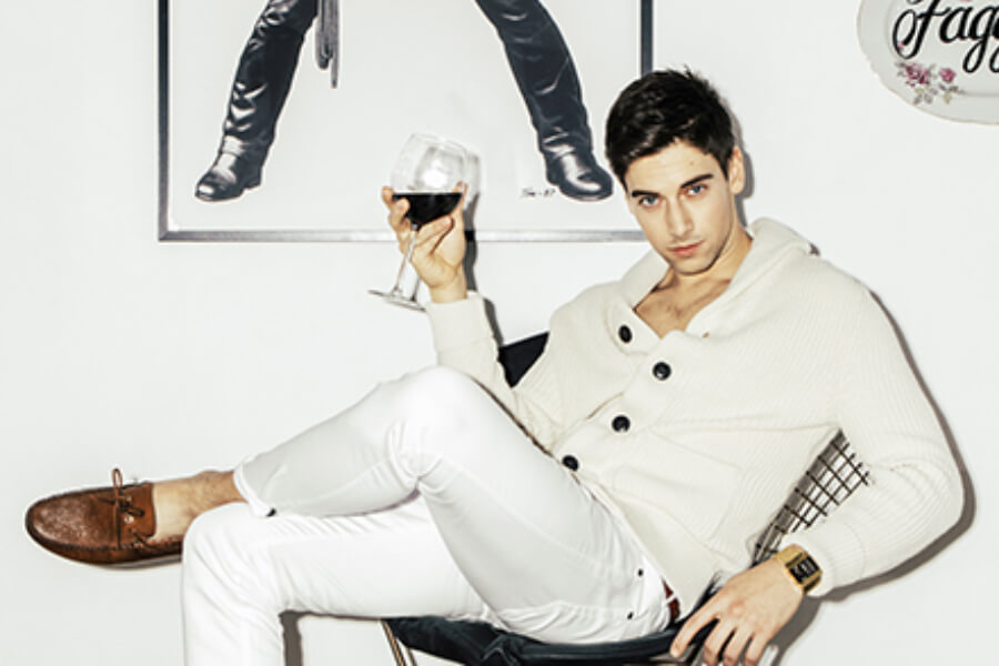 A man in a white sweater and white pants sits casually in a chair, holding a glass of red wine, with a poster of a dancer on the wall behind him.