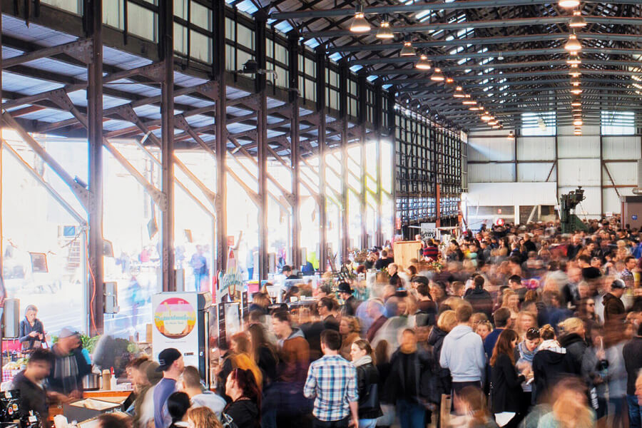 A busy indoor market scene filled with a large crowd, featuring stalls and natural light streaming through large windows.