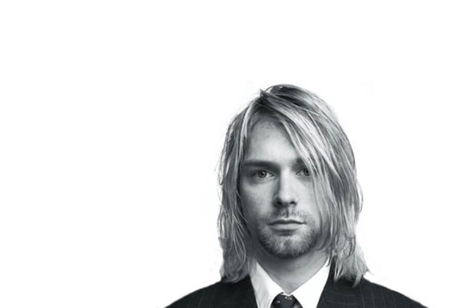 A young man with long, tousled hair wears a formal suit and tie, staring directly into the camera against a plain white background.
