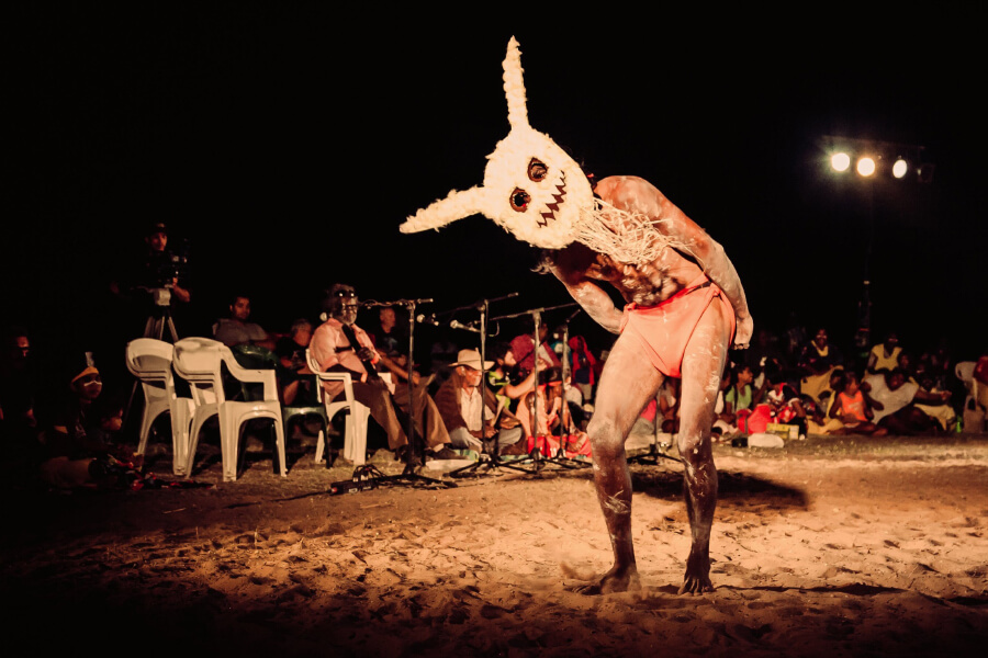 A performer wearing a whimsical mask and minimal clothing dances on a sandy stage, surrounded by an audience at night.