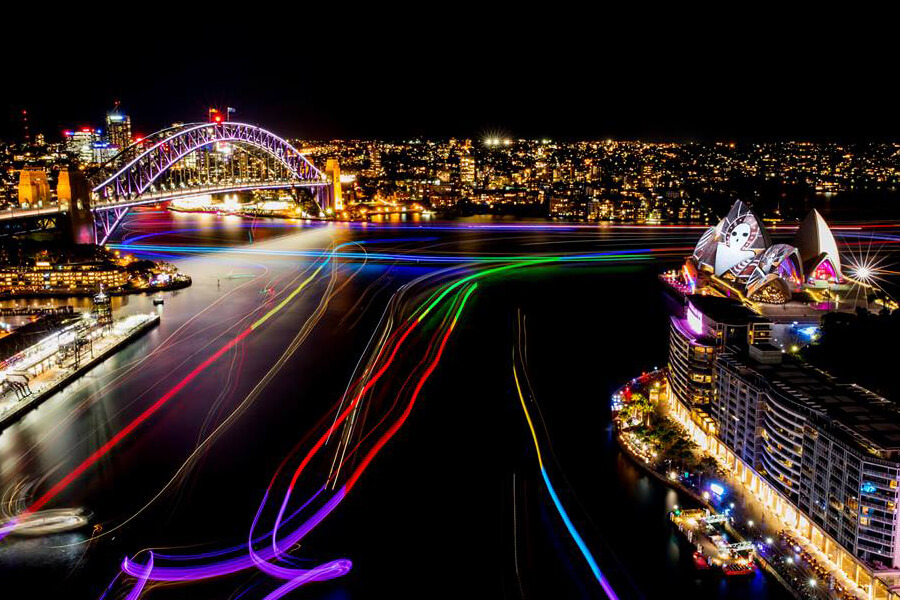 Sydney Harbour is illuminated at night, showcasing the Sydney Opera House and Harbour Bridge, with colorful light trails from boats on the water.