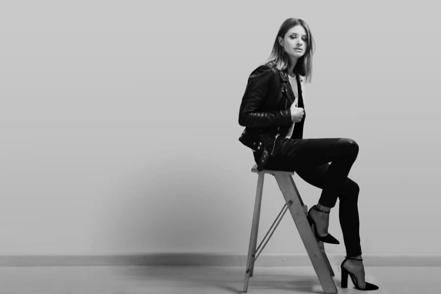 A woman in a black leather jacket and heeled shoes sits on a wooden stool against a light gray backdrop.