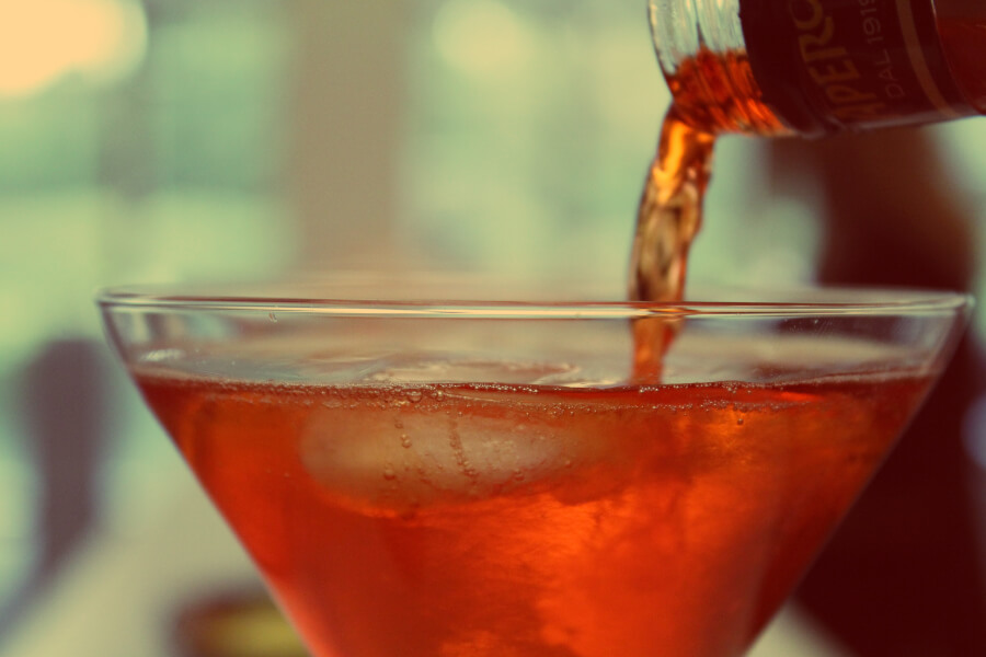 A close-up of a cocktail glass with ice, as a reddish-brown liquid is being poured into it.