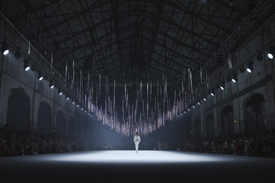 A runway model walks down a well-lit catwalk under hanging light fixtures in an industrial venue with an audience on either side.