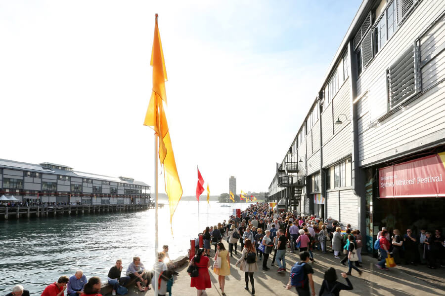 Crowds gather along a waterfront promenade lined with flags and buildings during a festival, with sunlight shining brightly.