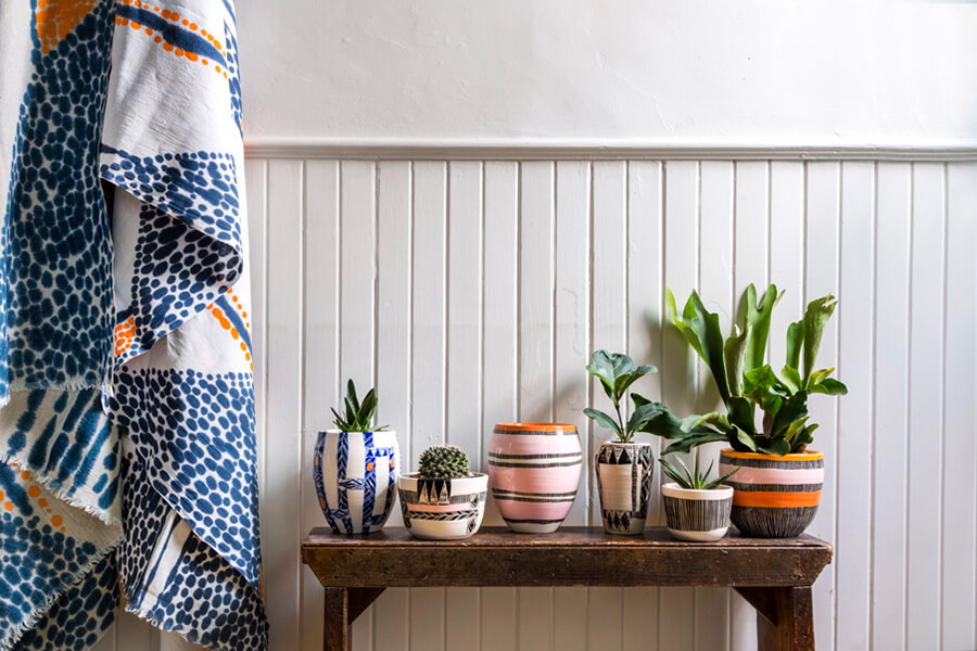 A wooden table displays several decorative plant pots of various colors and patterns, with green plants in each pot. A patterned blanket hangs beside the table against a white paneled wall.