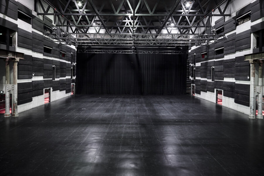 A spacious performance venue with high ceilings, black walls, and a polished floor, featuring a black curtain at the back.