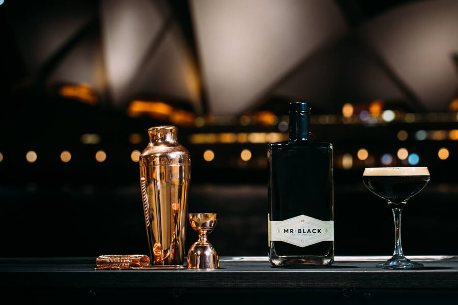 A black bottle labeled "Mr. Black" is placed on a table with a cocktail glass, a shaker, and a jigger, against a blurred evening backdrop.