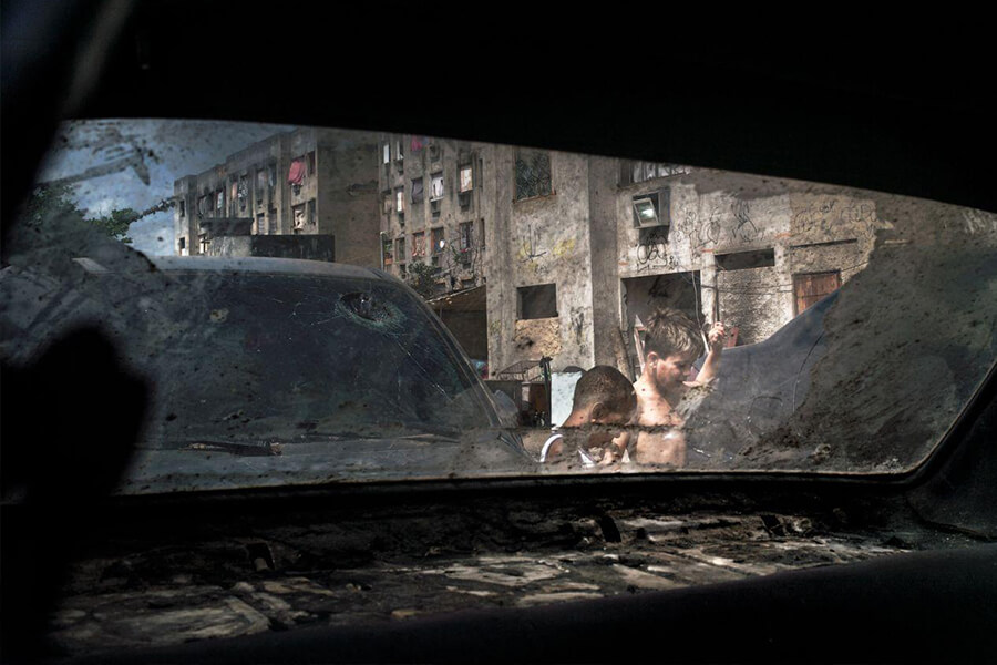 View through a dirty car window showing two children playing among abandoned vehicles and dilapidated buildings in the background.