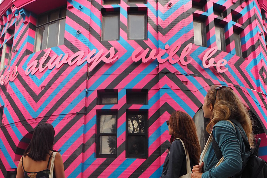 Three people observe a colorful, geometric mural on a building wall, featuring the phrase "always will be" in pink lettering.