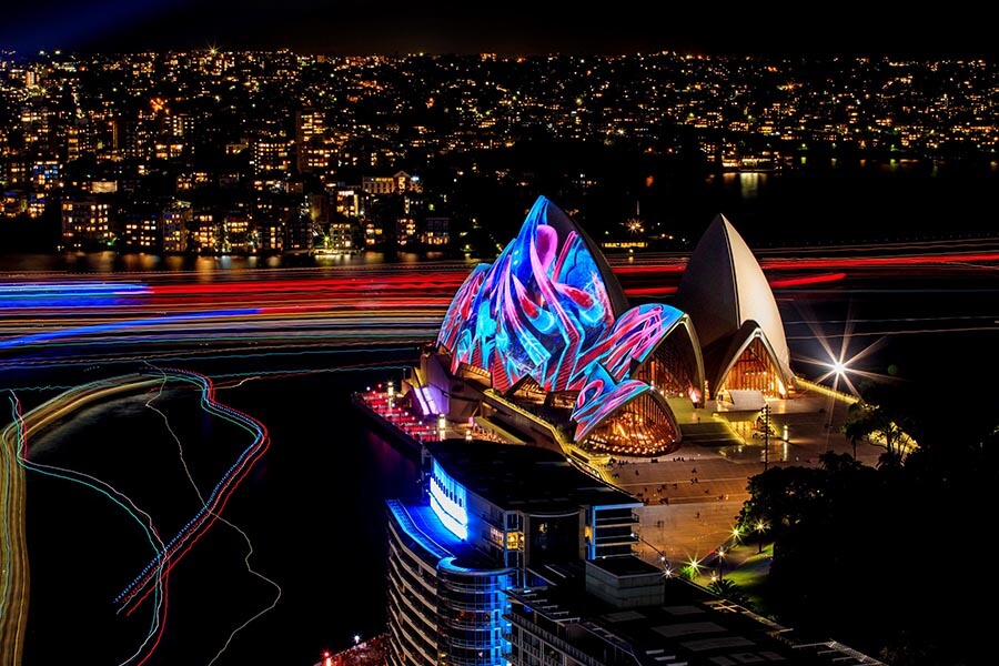 The Sydney Opera House is illuminated with colorful lights at night, with a vibrant city skyline in the background.
