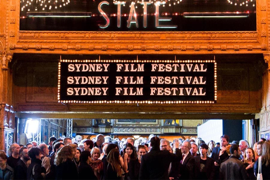 Crowd gathered outside a building with a marquee displaying "SYDNEY FILM FESTIVAL," featuring lights and ornate architecture.