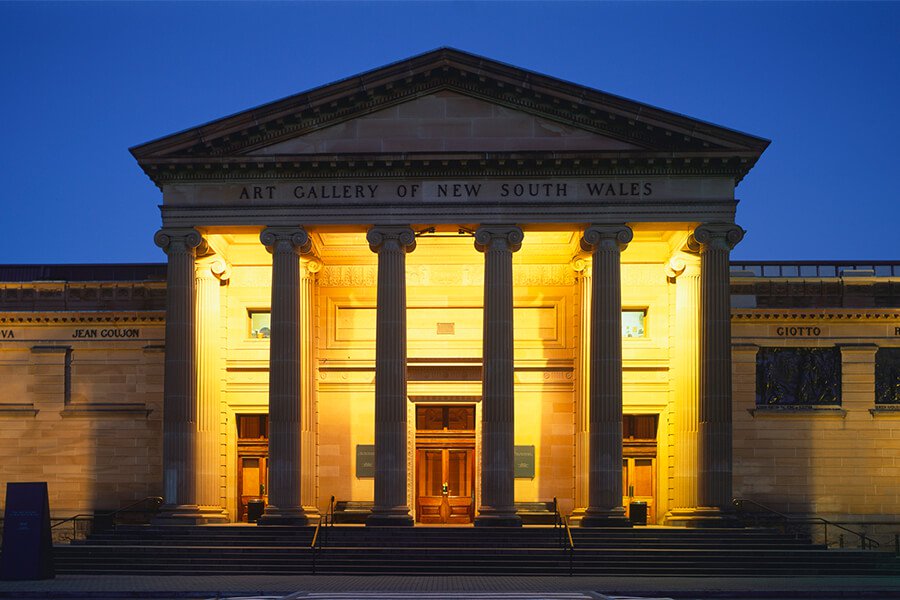 The front façade of the Art Gallery of New South Wales is illuminated at night, showcasing grand columns and entrances.