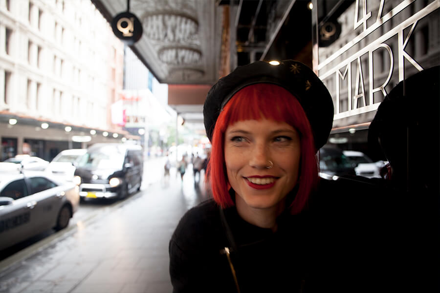 A smiling person with red hair and a black beret appears near a shop window, with city streets and cars in the background.