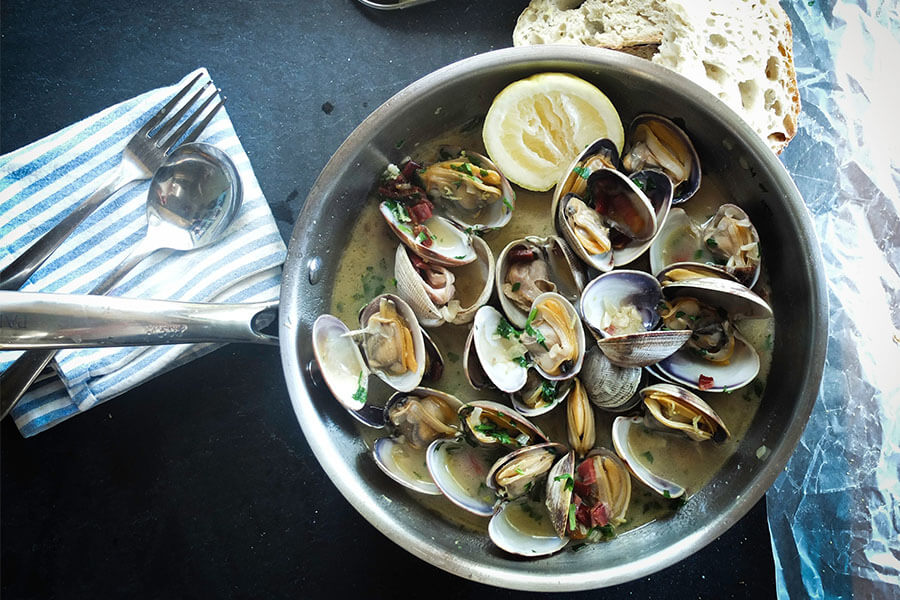 A pan filled with clams in broth, garnished with herbs and a lemon slice, alongside a slice of bread and utensils.