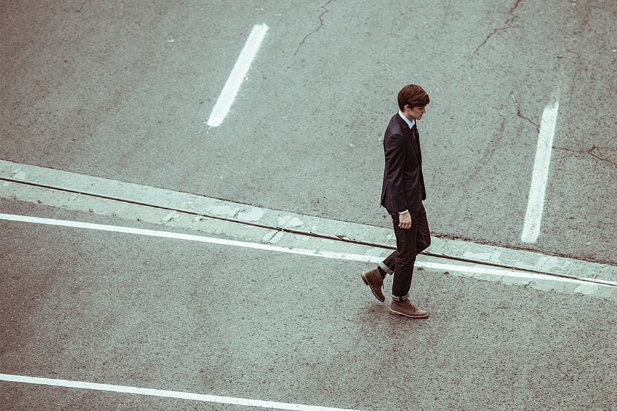 A man in a suit walks alone on an empty road, with white lane markings visible on the asphalt.