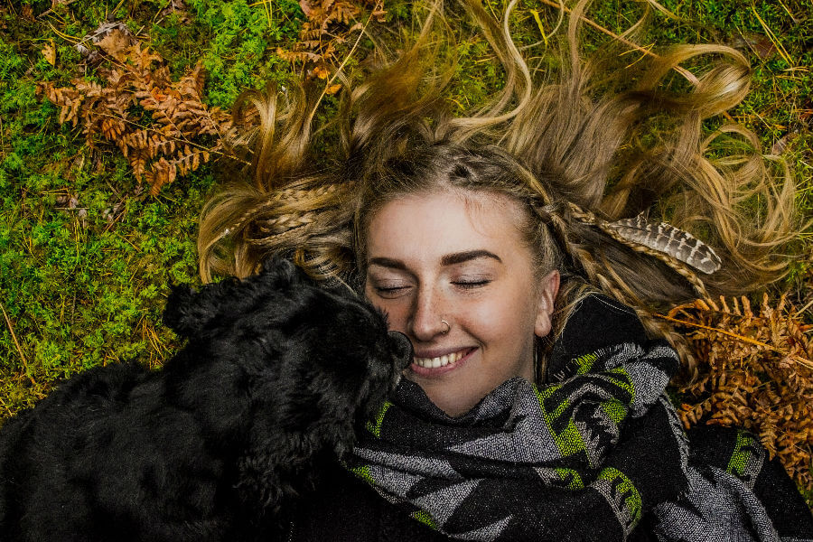 A smiling person with long hair lies on the ground, closely interacting with a black dog amidst foliage.