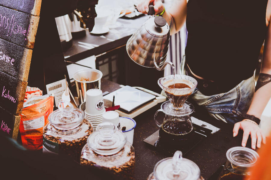 A person pours hot water from a kettle into a coffee dripper set over a glass carafe on a coffee counter. Jars of coffee beans and cups are visible.