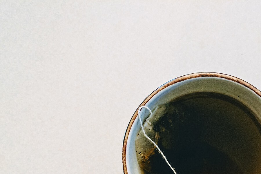 A close-up view of a mug filled with dark liquid, featuring a tea bag submerged in it. The background is a light, neutral color.