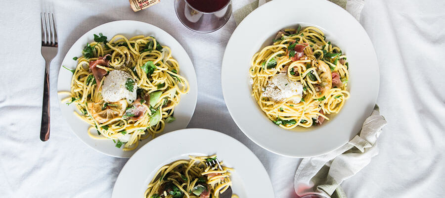 Three plates of pasta with vegetables and a poached egg, accompanied by a glass of red wine and a fork on a white tablecloth.