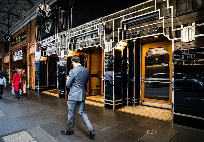 A man in a suit walks past an art-deco styled building with multiple entrance doors, including one labeled "SCREENING ROOM."