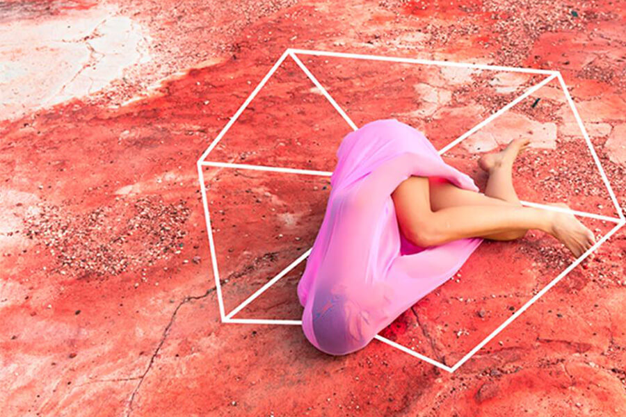 A person in a pink garment is curled on a red, textured surface, inside a geometric outline.
