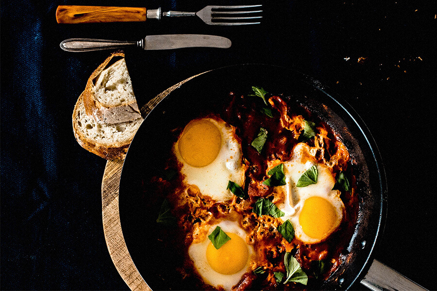 A skillet with three eggs cooked in a red sauce, topped with green basil, beside a slice of bread and cutlery.