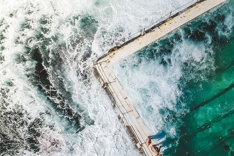 Aerial view of a pool at the ocean's edge, surrounded by vibrant waves and foam, with people visible on the edge.