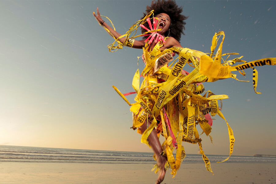 A person jumps joyfully on the beach, wearing a dress made of yellow caution tape, with a sunset in the background.